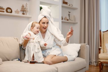 Genç anne ve küçük kız havluya sarılı sabahlıklar giyiyorlar. Kanepede oturan kadın ve kız. Kadın ve kız küçük aynaya bakıyor..