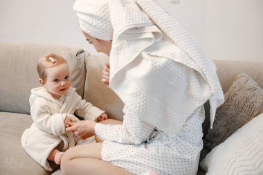 Genç anne ve küçük kız havluya sarılı sabahlıklar giyiyorlar. Kanepede oturan kadın ve kız. Kızın saçında toka var..