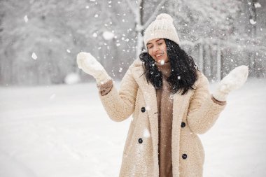Esmer kadın ormanda durup poz veriyor. Kadın karı gördüğüne sevindi. Kışlık kıyafetler giyen ve eldivenli şapka takan bir kız..