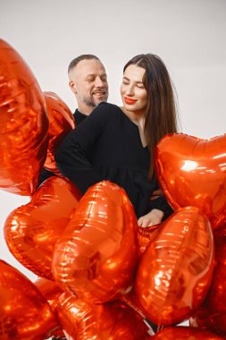 Yıldönümünde fotoğraf çektirmek için poz veren romantik bir çift. Adamın karısını kucakladığı ve elinde kalp şeklinde kırmızı balonlar tuttuğu bir stüdyo fotoğrafı. Esmer kadın ve siyah elbiseli sakallı adam.