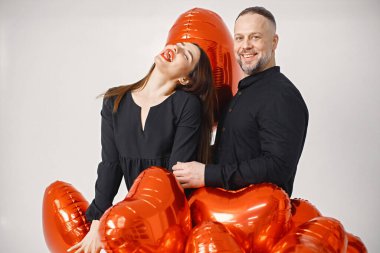 Yıldönümünde fotoğraf çektirmek için poz veren romantik bir çift. Adamın karısını kucakladığı ve elinde kalp şeklinde kırmızı balonlar tuttuğu bir stüdyo fotoğrafı. Esmer kadın ve siyah elbiseli sakallı adam.