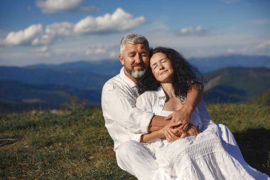 Dağlarda kıdemli adam ve kadın. Günbatımında aşık olan yetişkin bir çift. Beyaz gömlekli adam. Gökyüzü arka planında oturan insanlar.