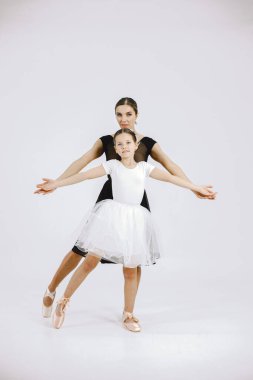 Little girl ballerina with her mother on white background. Brunette girl wearing white ballet suit and adult woman black dress. Two female dancer dancing together in a pointe shoes.