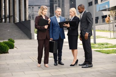 Modern ofis binasının önünde kendinden emin iş adamları konuşuyor. Belgelere bakan siyah ve beyaz erkekler ve sarışın kadınlar. Bankacılık ve finans piyasası kavramı.