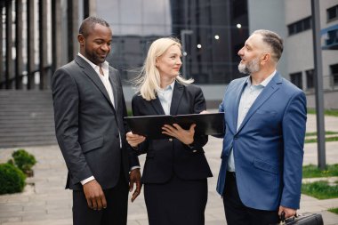 Modern ofis binasının önünde kendinden emin iş adamları konuşuyor. Belgelere bakan siyah ve beyaz erkekler ve sarışın kadınlar. Bankacılık ve finans piyasası kavramı.