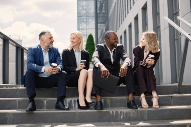 Modern ofis binasının önündeki merdivenlerde oturan kendine güvenen iş adamları. Erkekler ve kadınlar konuşuyor ve ellerinde hazır kahve var. Bankacılık ve finans piyasası kavramı.