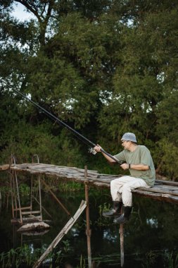 Yazın göl kenarında olta atan bir adam. Bir yaya köprüsünde oturan balıkçı. Haki elbiseli ve şapkalı bir adam..
