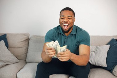Black man sitting on a coach and holding a cash. Man wearing blue t-shirt. Man posing for a photo and looking very happy.