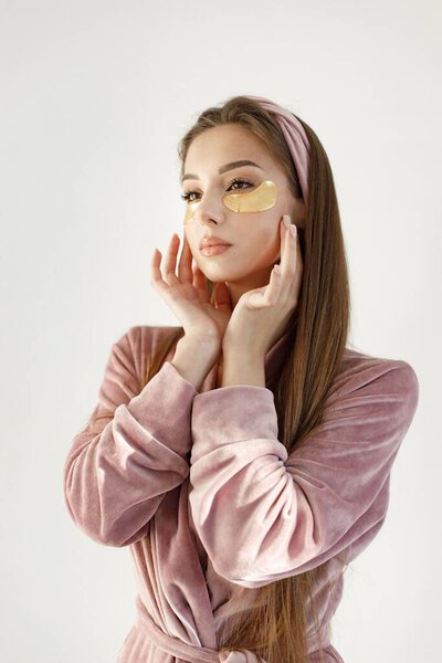 Young woman applying a face patches. Brunette woman standing on a white backgroung. Girl wearing rose pajamas and headband.