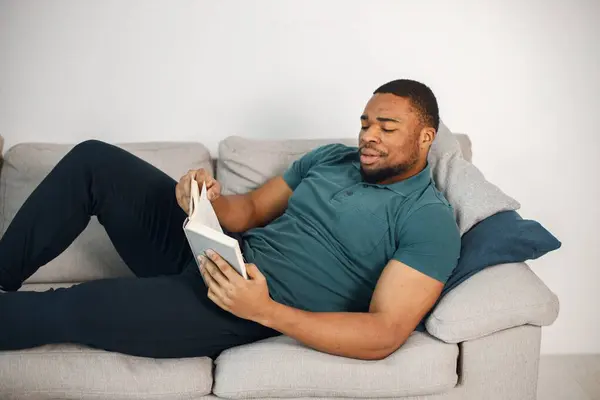Black man lay on a coach with a book. Man wearing blue t-shirt. Man reading a book at home on a coach.