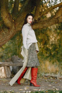 Beyaz kadın dışarıda duruyor ve bir fotoğraf için poz veriyor. Gri şapkalı, ceketli ve etekli bir kadın. Kadının kırmızı dudakları var..