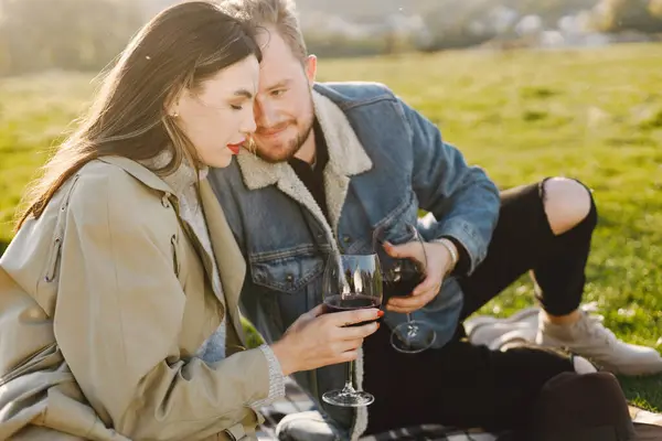 Romantic couple in fashion clothes sitting on a nature on a picnic rug. Man wearing jacket and woman coat. Woman and man drinking a wine.