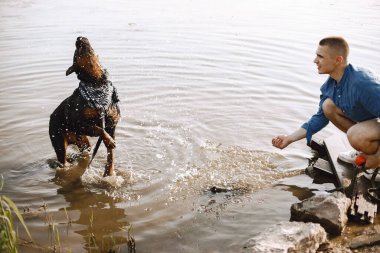 Handsome young male in casual outfit playing with cute dog while sitting near the lake. Boy wearing blue shirt and white jeans shorts. Dog has a white and black scarf on his neck.