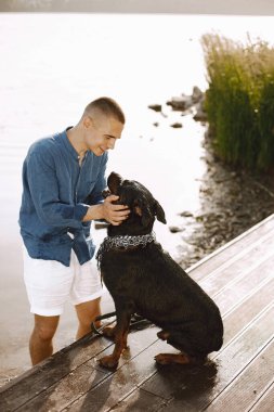 Handsome young male in casual outfit playing with cute dog while standing near the lake. Boy wearing blue shirt and white jeans shorts. Dog has a white and black scarf on his neck.