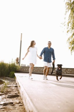 Romantic couple boyfriend and girlfriend walking with rotweiller dog near the lake together. Man and woman looking at each other. Man wearing blue shirt and woman white dress.