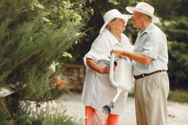 Adult couple in a summer garden. Handsome senior in a white shirt. Woman in a hat. Family watering.