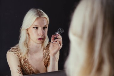 Young blond man holding brushes and doing make up on his face. Man wearing fashionable bright golden t-shirt. Man looking in a studio mirror.
