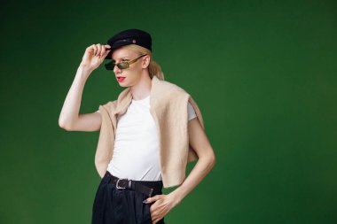 Young blond man with make up on his face and sunglasses. Man wearing fashionable white t-shirt and black cap. Man isolated on green background.