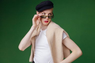 Young blond man with make up on his face and sunglasses. Man wearing fashionable white t-shirt and black cap. Man isolated on green background.