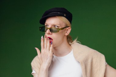 Young blond man with make up on his face and sunglasses. Man wearing fashionable white t-shirt and black cap. Man isolated on green background.