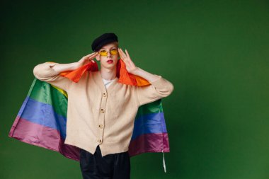 Young blond man with make up on his face wearing sunglasses. Man wearing fashionable beige sweater and black cap. Man isolated on green background and holding rainbow flag.