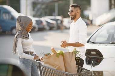 Afrikalı Arap çift, markette elektrikli arabanın yanında duruyor. Elektrikli benzin istasyonunda elektrikli araba şarj ediliyor.