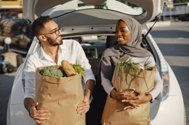 Alışverişten sonra eğlenen mutlu çift. Taze ve sağlıklı yiyeceklerle dolu bir arabanın bagajında oturuyor. Afrikalı Müslüman kadın ve Arap adam.