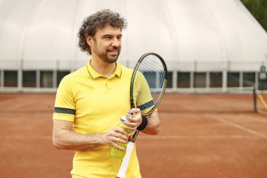 Portrait of tennis player man, front view. Man standing on a court and holding a racquet. Man wearing yellow t-shirt. clipart