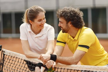 Curly man and woman tennis players standing on open summer court with rackets in hands. Couple friendly chatting after the match while leaning on a net. Man wearing yellow t-shirt and woman white one.