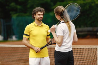 Curly man and woman tennis players standing on open summer court with rackets in hands. Couple friendly chatting before the match. Man wearing yellow t-shirt and woman white one.