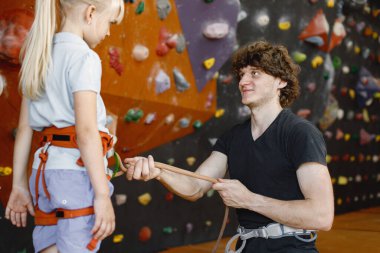 Eğitmen, küçük bir kızın içerideki kaya tırmanış merkezinde emniyet kemeri takmasına yardım ediyor. Küçük kız, çocuk eğlence merkezinde kaya tırmanışı yapıyor. Sarışın Kafkasyalı