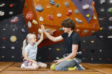 Erkek eğitmen, spor salonunda küçük bir kızla konuşuyor. Duvardaki yapay taşları gösteren güvenlik gereksinimlerini anlatıyor. Küçük kız, çocukların eğlencesinde kaya tırmanışı yapıyor.
