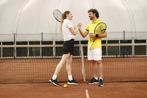 Curly man and woman tennis players standing on open summer court with rackets and balls in hands. Couple friendly shaking hands at court before the match. Man wearing yellow t-shirt and woman the