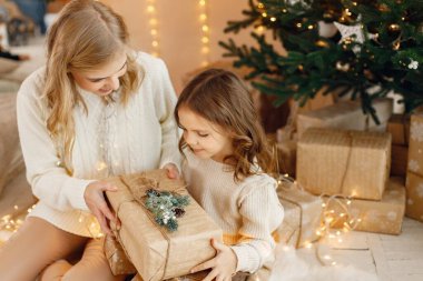 Noel 'i kutlayan ve bir hediye kutusu tutan küçük bir kızın portresi. Kız ve annesi Noel ağacının yanında oturup poz veriyorlar. Bej örgülü giysiler giyen bir aile..