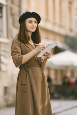 Kadın ressam şehir merkezinde dururken resim yapıyor. Haki elbisesi ve siyah şapkası olan bir kadın. Yazın sokakta çizim yapan bir kız..