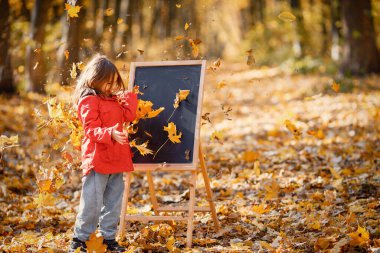 Photo of little girl walking in autumn forest. Girl playing near easel and holding a yellow leaves. Cute girl wearing red jacket.