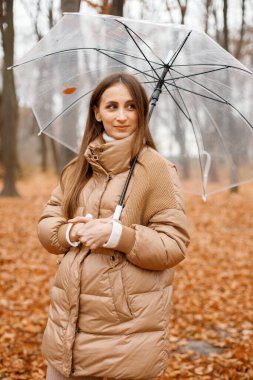 Young woman standing in autumn forest. Brunette woman holding a transparent umbrella. Girl wearing fashion brown jacket.