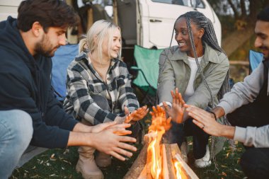 Çok ırklı arkadaşlar şehir dışında piknik yapıyorlar. Ateş yaktılar ve sıcak elleriyle oturdular. Avrupa, siyah, Pakistan ırkı. Minibüsle yolculuk..