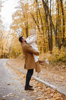 Young man and woman walking outside wearing beige coats. Blond woman and brunette man in autumn forest. Romantic couple kissing.