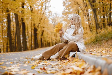 Young woman sitting on a curb in autumn forest. Blonde woman holding a yellow leaves. Girl wearing beige coat and brown trousers.