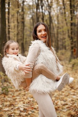 Young woman with little girl standing in autumn forest. Brunette woman holding her daughter piggyback. Girl wearing beige sweater and mother wearing white clothes.
