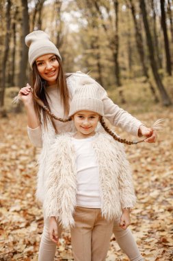 Young woman with little girl walking in autumn forest. Brunette woman play with her daughter. Girl wearing beige sweater and mother wearing white clothes.