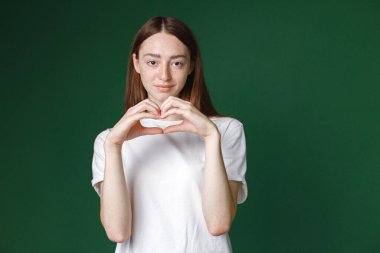 Yeşil arka planda izole edilmiş beyaz bir kızın kapalı bir portresi. Beyaz tişörtlü, fotoğraf için poz veren ve elleriyle kalbi gösteren güzel bir kadının portresi. Genç bir kızın stüdyo fotoğrafı..