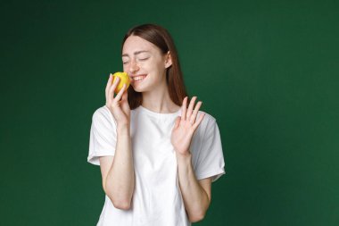 Yeşil arka planda izole edilmiş beyaz bir kızın kapalı bir portresi. Bir fotoğraf için poz veren beyaz tişörtlü güzel bir kadının portresi. Limon tutan genç bir kızın stüdyo fotoğrafı..