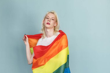Yüzünde makyaj ve gökkuşağı bayrağı olan genç sarışın bir adam fotoğraf için poz veriyor. Beyaz tişörtlü ve kırmızı dudaklı bir adam. Mavi arkaplanda izole edilmiş bir adam.