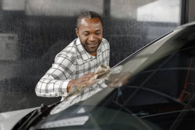 Man washing luxury car on a car wash with a rag. Black man wiping his car. Man wearing plaid shirt, smiling.