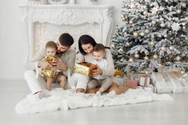 Anne, baba ve iki oğlu Noel ağacının yanında oturuyor. Heyecanlı aile Noel hediyelerini evde açıyor. Beyaz elbiseli kadın, erkek ve iki çocuk..