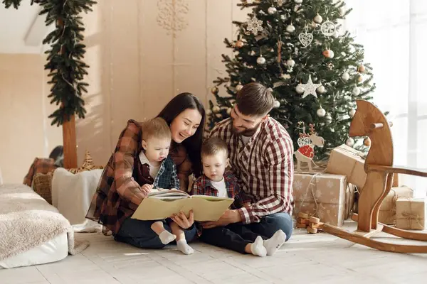 Anne, baba ve iki oğlu Noel ağacının yanında oturuyor. Ailesi kitap okuyan çocuklar. Kadın, erkek ve iki çocuk ekose gömlek giyiyor..