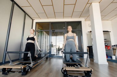 Özel bir ekipman üzerinde egzersiz yapan iki hamile kadın. Sarışın ve esmer kadınlar spor salonunda çalışıyor. Gri ve siyah spor ayakkabı giyen kadınlar..