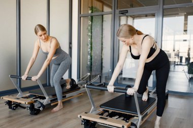 Özel bir ekipman üzerinde egzersiz yapan iki hamile kadın. Sarışın ve esmer kadınlar spor salonunda çalışıyor. Gri ve siyah spor ayakkabı giyen kadınlar..